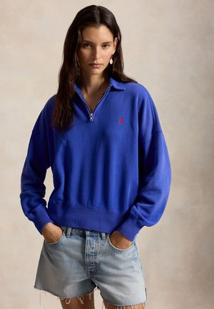 Pikkade tumejate juustega naine, kes kannab sinist kvartstõmblukuga pulloverit ja heledate teksapükstega lühikesi sorte, käed taskutes, beežist taustast ees.