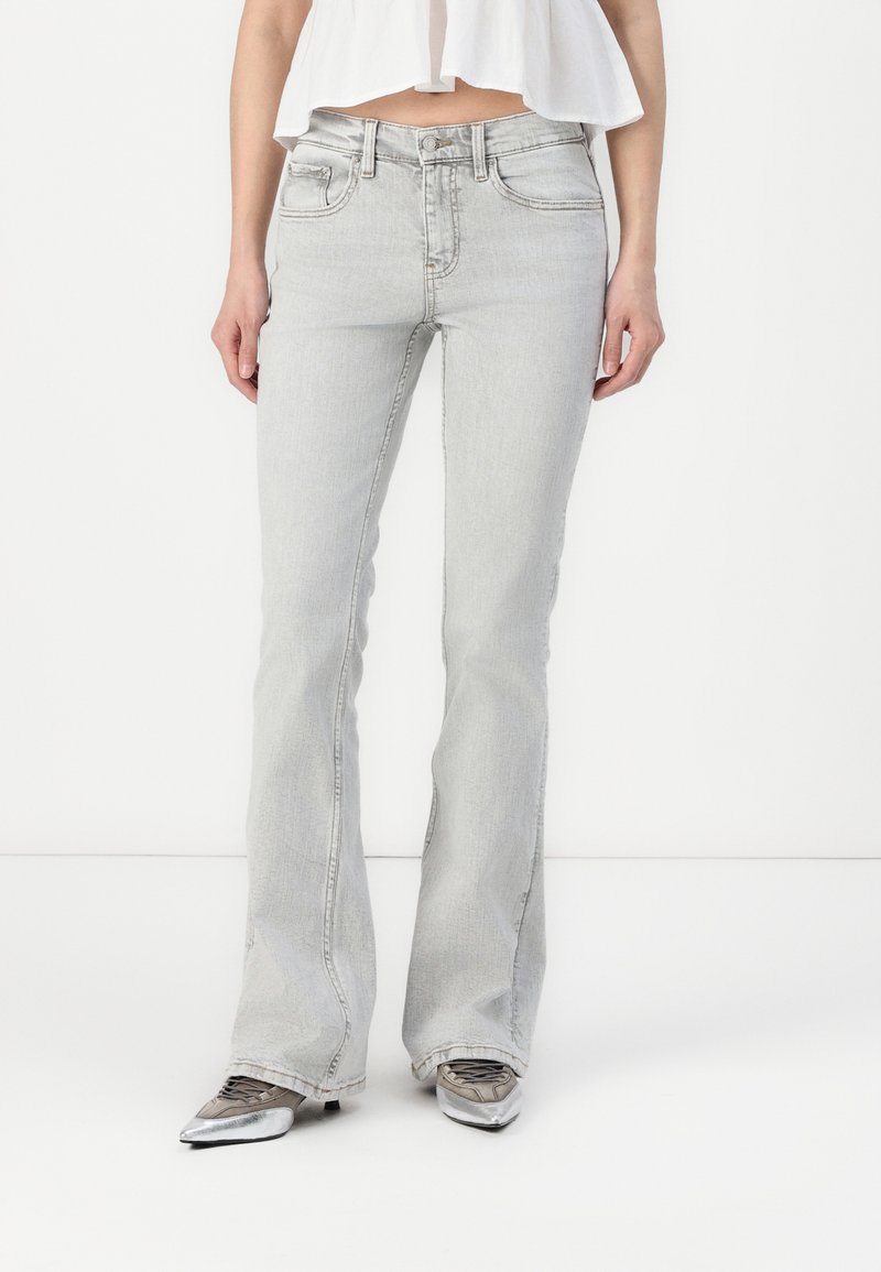 Jeans de campana gris claro con un ligero estiramiento, que presentan bolsillos frontales y detalles de costura sutiles. Llevados con zapatos plateados de punta.