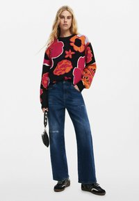Maglione nero con vivaci motivi floreali in rosso, arancione e viola; abbinato a jeans blu a vita alta e sneakers nere.