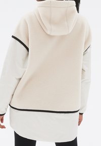 Krämfärgad fleecejacka med huva, som har ett slätt vitt tyg som underlager och svarta detaljer. Texturerad yta och förlängd bakre fåll.