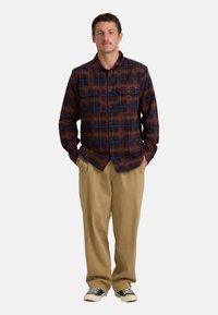 Ruitjes flanellen overhemd in donkerrode en blauwe tinten, met twee borstzakken en een knoopsluiting, gecombineerd met beige broeken en zwarte sneakers.