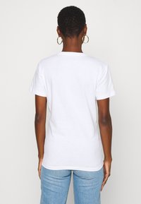 Camisa de algodón blanca de manga corta, con un escote redondeado y un corte relajado, vista desde detrás sobre un fondo claro y liso.