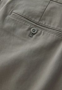 Gros plan sur un pantalon en tissu gris montrant un seul bouton, une boutonnière et une poche arrière horizontale avec des détails de couture.