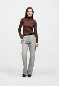 Pruun pikkade varrukatega särk võltskaelusega ja sinise logoga, mida kombineeritakse heledate hallide sirge lõikega teksade ning mustade teravate ninaga kontsakingadega. Lihtne taust.