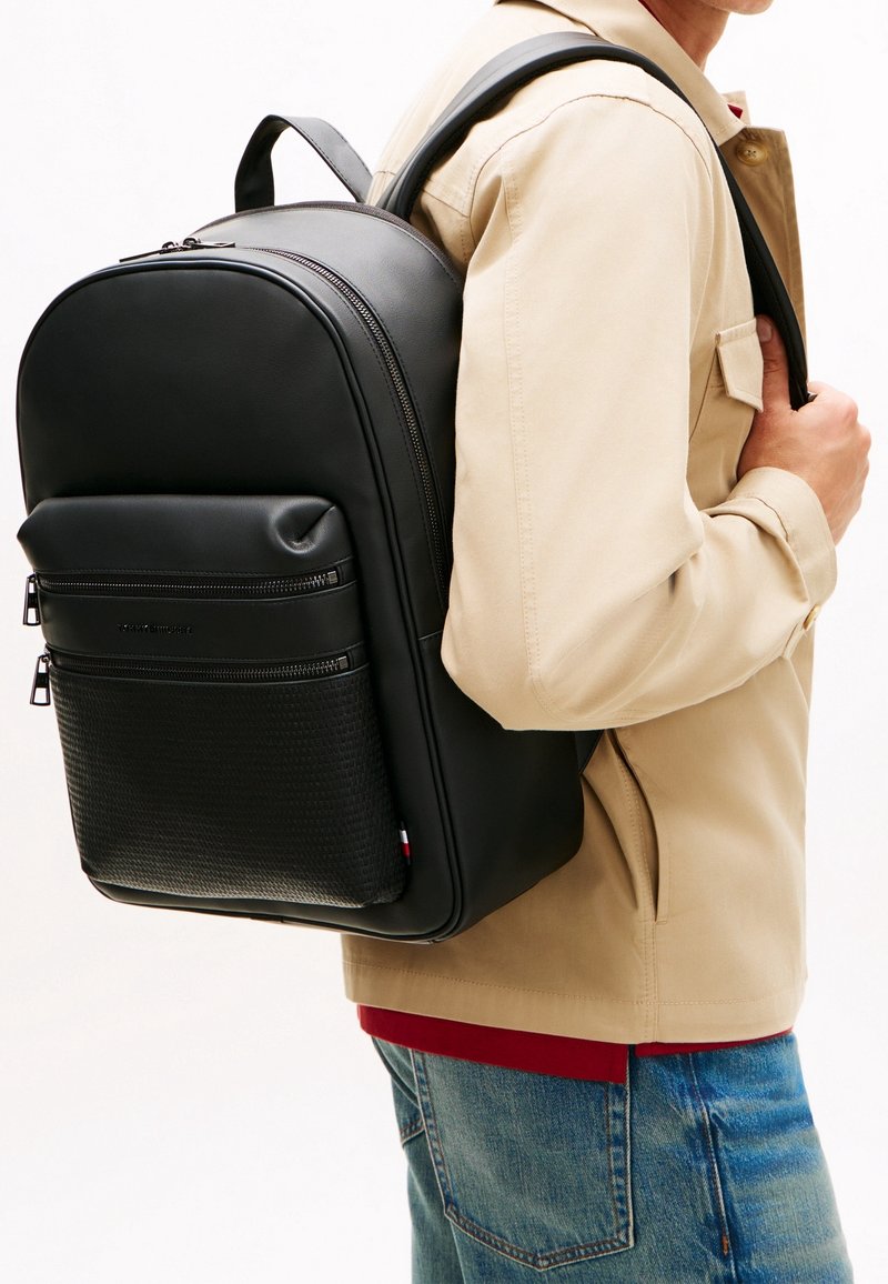 Personne portant une veste beige et un jean bleu, avec un sac à dos en cuir noir comportant deux poches avant zippées.