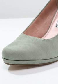 Chaussure à talon haut en daim vert clair avec bout arrondi, plateforme discrète et doublure intérieure rose pâle sur une surface blanche.