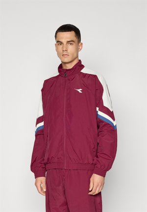 Mand iført en vinrød sportsjakke med hvide og blå striber på ærmerne, stående foran en ensartet lys baggrund.