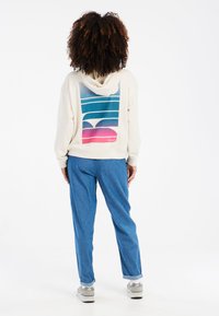 Felpa con cappuccio color crema con grafica colorata sul retro; abbinata a pantaloni blu, che mostrano una vestibilità rilassata. Sneakers bianche visibili.