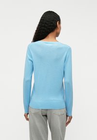 Világoskék, puha anyagból készült pulóver, hosszú ujjal és lekerekített nyakkivágással. Testhez álló fazonnal és bordázott aljjal rendelkezik, szürke nadrággal viselve.