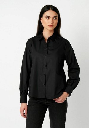 Frau mit langen dunklen Haaren, die ein schwarzes Hemd mit Knöpfen und schwarze Hose trägt, steht mit einer Hand in der Tasche und schaut zur Seite.