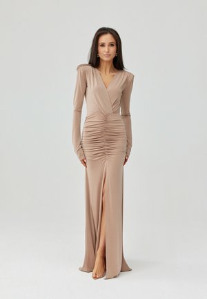 Vrouw draagt een lange, beige jurk met plooien, lange mouwen en een split aan de voorkant, staand tegen een effen lichtgrijze achtergrond.