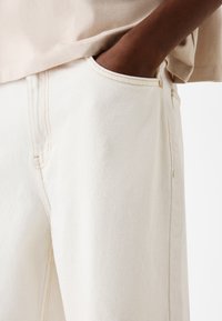 Beige puuvillajegginsit, joissa on teksturoitu pinta, näkyvät saumat ja sivutasku. Kankaassa on hieman painoa.