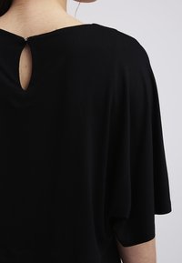 Černý top s volnějším střihem, krátkými rukávy a kulatým výřezem na zádech s dekorativním detailem knoflíku. Měkká textilní struktura.