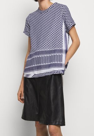 Osoba nosząca krótki, wzorzysty niebiesko-biały top oraz czarne, skórzane szorty do kolan, stojąca z jedną ręką w kieszeni.