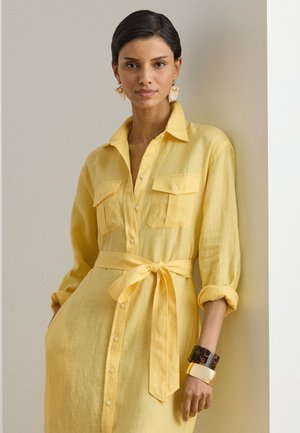 Kobieta ubrana w żółtą sukienkę zapinaną na guziki, z przednimi kieszeniami, przewiązanym paskiem, podwiniętymi rękawami, kolczykami muszelkowymi i dwoma bransoletkami, stojąca na tle białej ściany.