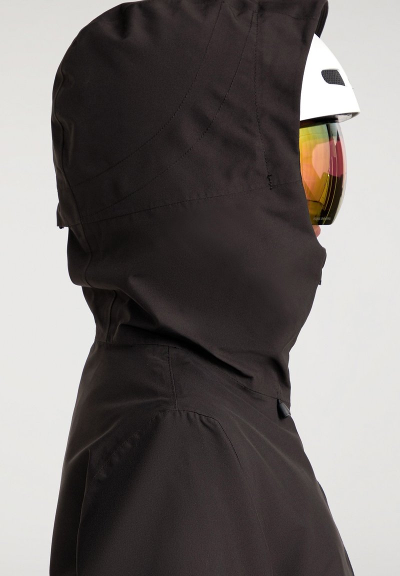 Chaquetón negro impermeable con un alto cuello, que presenta una textura suave y un diseño ajustado. Se puede ver un casco blanco con visor ahumado.