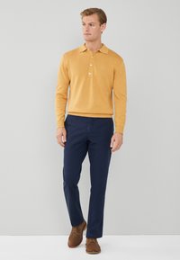 Polo giallo senape a maniche lunghe con tre bottoni, abbinato a pantaloni navy. Scarpe in suede marroni completano l'outfit. Sfondo neutro.
