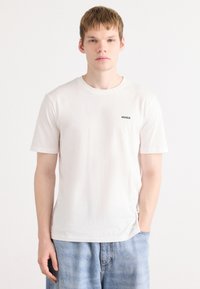 Hvit bomulls t-skjorte med korte ermer, med en liten svart "HUGO"-logo på brystet. Kombinert med lyseblå denimshorts.