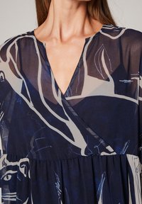 Marineblaues Kleid aus durchscheinendem Stoff mit geometrischen weißen und blauen Mustern, V-Ausschnitt und gerafftem Taillenbereich für eine fließende Silhouette.