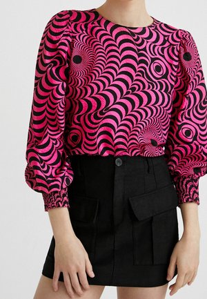 Femme portant un chemisier à manches longues rose et noir à motif illusion d'optique avec manches bouffantes et une mini-jupe noire avec poches avant.
