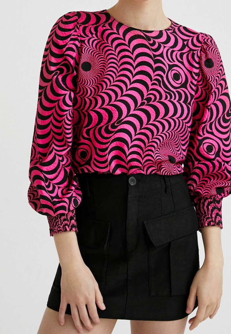Femme portant un chemisier à manches longues rose et noir à motif illusion d'optique avec manches bouffantes et une mini-jupe noire avec poches avant.