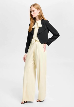 Vrouw met lang rood haar draagt een zwarte cropped blazer, crème blouse met strik, crème wijde broek en zwarte open-teen hakken, staande zijwaarts.