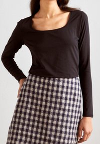 Kvinna som bär en svart långärmad topp med en asymmetrisk halsringning och en knälång svart och vit rutigt kjol.