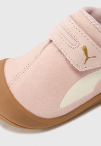 Chaussure en daim rose avec une semelle en caoutchouc, équipée d'une sangle velcro et d'une garniture beige clair. Logo doré sur le côté. Texture lisse dans l'ensemble.
