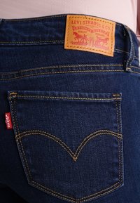 Jeans de mezclilla de un azul oscuro con un parche de cuero visible en la cintura que presenta el logo de Levi Strauss & Co. y detalles de costura dorada en los bolsillos.