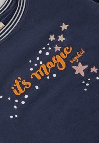 Tessuto blu navy con testo ricamato arancione "è magia" circondato da stelle beige e rosa e pallini bianchi, colletto a righe visibile.