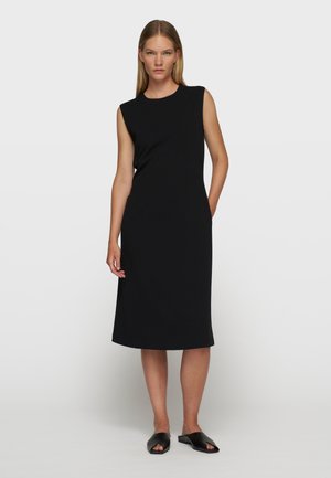 Donna che indossa un abito nero senza maniche al ginocchio con spalline imbottite, in piedi con una mano in tasca e scarpe nere aperte sulle dita.