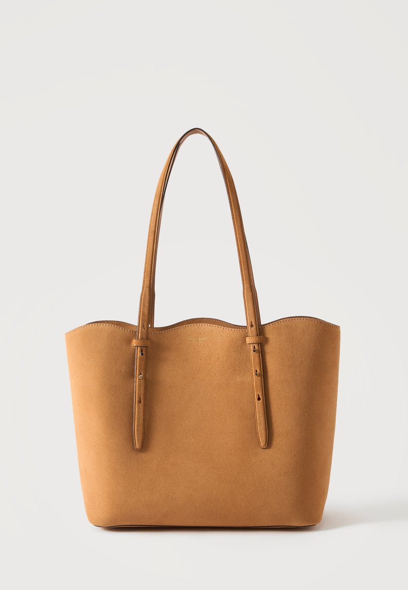 Sac cabas en daim beige avec deux bretelles réglables et un bord supérieur légèrement incurvé sur fond uni.