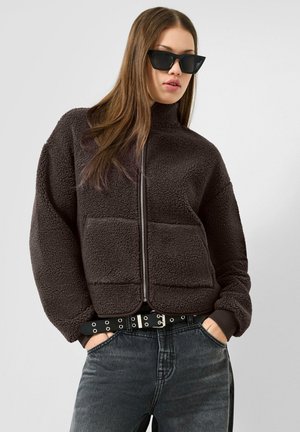 Donna che indossa una giacca nera morbida con zip, jeans scuri, cintura nera con occhielli argentati e occhiali da sole neri a occhi di gatto, con le mani nelle tasche.