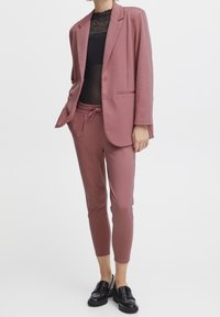 Rosa kavaj med två knappar och framfickor, matchad med joggerbyxor i samma färg och en genomskinlig svart topp. Svarta loafers fulländar looken.