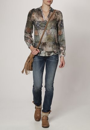 Nainen, jolla on kuvioitu pitkähiha paita, siniset farkut, vaaleanruskeat nilkkurit ja vaaleanruskea hapsullinen olkalaukku, kävelee yksiväristä taustaa vasten.