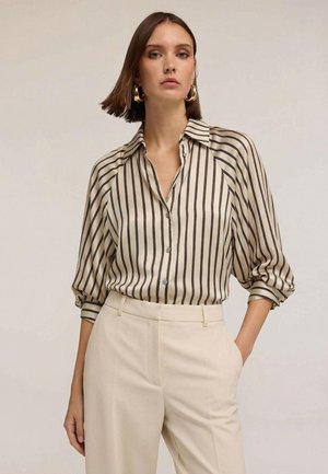 Camicia beige a righe con bottoni e maniche arrotolate, abbinata a pantaloni crema, caratterizzata da una vestibilità comoda e una texture liscia.