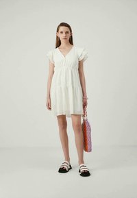 Jovem mulher vestindo um vestido de renda branco e sandálias brancas, segurando uma bolsa de palha trançada multicolorida, de pé contra um fundo simples.