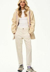 Kobieta o długich blond włosach, ubrana w beżową kurtkę, białą koszulę, kremowe spodnie oraz kolorowe trampki, stojąca z rękami w kieszeniach.