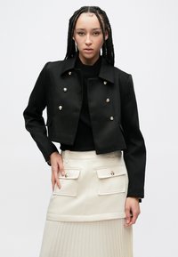 Giacca corta nera con bottoni dorati, abbinata a una gonna plissettata color crema con tasche frontali e una texture liscia.