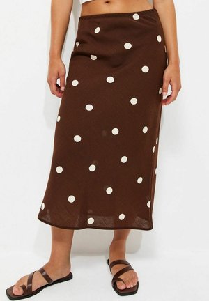 Jupe midi marron à pois blancs portée par une personne debout, associée à des sandales plates marron à brides.