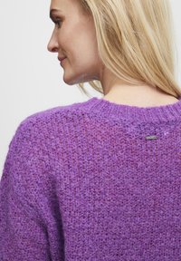 Pull en tricot violet avec une texture lâche et un col rond. Présente une petite étiquette à l'arrière, soulignant le design décontracté.