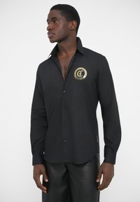 Camiseta negra de manga larga con un gran logo dorado en el pecho, con cuello abotonado y textura de tela suave.