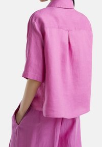 Růžová košile s krátkým rukávem z lehkého lnu, s záhybem na zádech a volnějším střihem, s bočními kapsami a čistými švy.