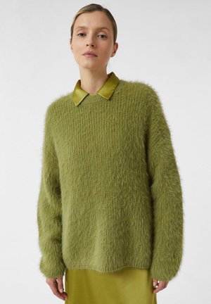 Frau trägt einen flauschigen olivgrünen Pullover über einem passenden Satinhemd mit Kragen und Rock, steht vor einem einfarbigen hellen Hintergrund.