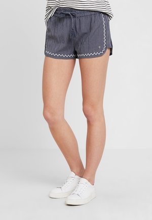 Personne portant un short bleu marine à fines rayures avec un bord en zigzag blanc et des baskets blanches, debout sur un fond uni clair.