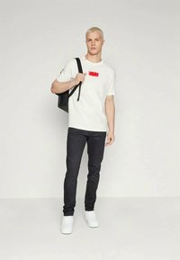Maglietta bianca con grafica rossa, jeans neri e sneakers bianche. Il modello porta uno zaino nero; lo sfondo è semplice e chiaro.