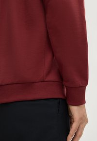 Felpa borgogna con polsini e orlo a coste. Tessuto liscio con una vestibilità aderente, abbinata a pantaloni scuri.
