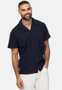 Camisa de manga corta en color navy con cuello de campamento, cierre de botones y tejido suave. Se lleva con pantalones de color claro, que presentan un sutil detalle de costura.