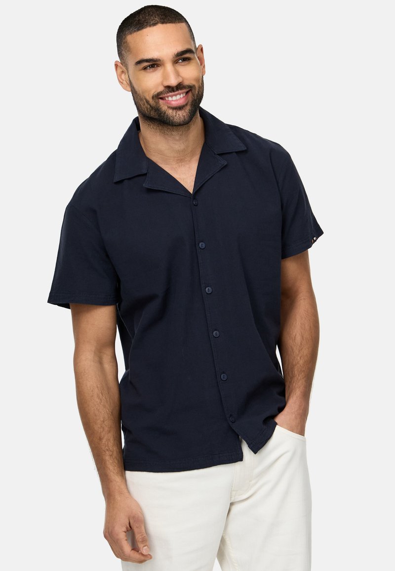 Camisa de manga corta en color navy con cuello de campamento, cierre de botones y tejido suave. Se lleva con pantalones de color claro, que presentan un sutil detalle de costura.