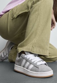 Baskets en suède gris avec des accents et des lacets blancs, posées sur un pantalon large olive, sur une surface grise.